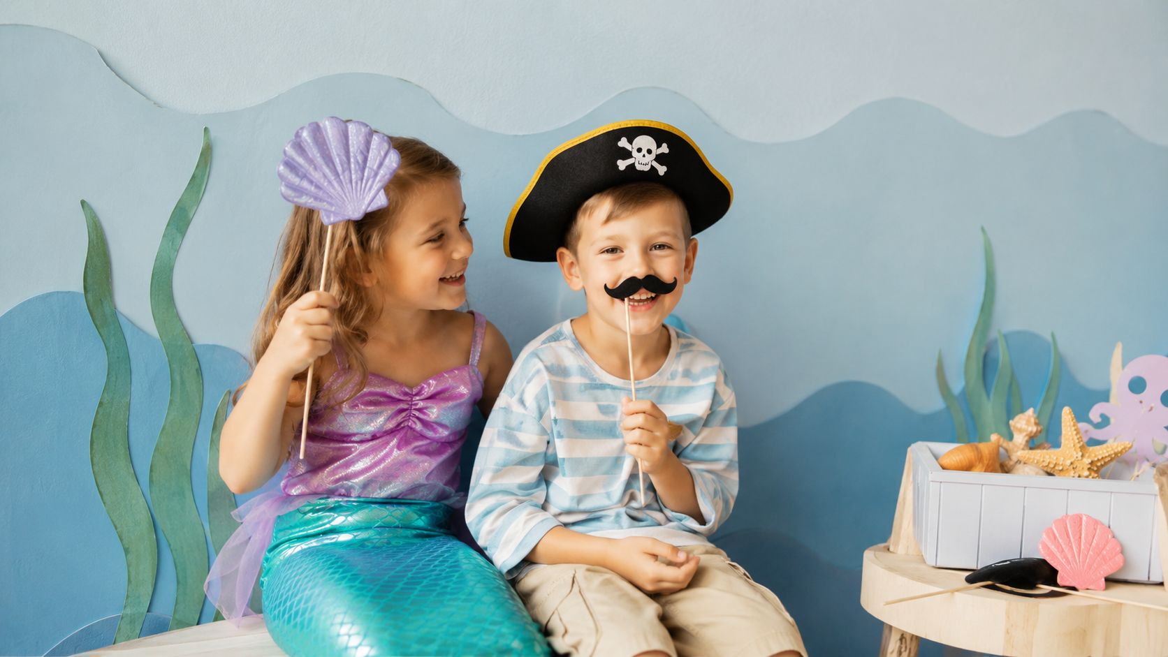 Cheerful children dressed as a mermaid and a pirate celebrate a birthday during a photo session against a decorated sea-themed backdrop.